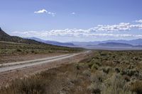 dirt road through dry brush and grasses near mountains with clouds above them and distant sky with sparse vegetation