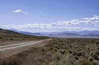 dirt road through dry brush and grasses near mountains with clouds above them and distant sky with sparse vegetation