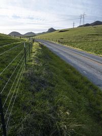 a grassy hill with electric wire fence in the foreground and distant country side with a road in the distance