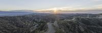 a winding road with mountains and the sun setting on the horizon in background, during a hazy day