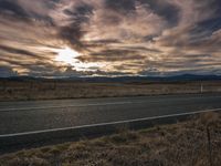 a road stretches into the sunset and some clouds above it, along with an empty highway running through grass, and tall, dry grass