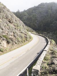 the car is driving on the curved road beside the hill and trees in the mountains