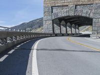 a tunnel leading to a highway near mountains in a scenic setting with the road's railings pointing out