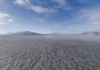 the lone man is walking across the barren area in the distance are mountains, trees, and a hazy blue sky