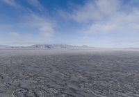 the lone man is walking across the barren area in the distance are mountains, trees, and a hazy blue sky