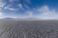 the lone man is walking across the barren area in the distance are mountains, trees, and a hazy blue sky