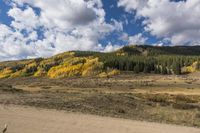 the road passes a sandy hill and forest filled with golden fall leaves as a person rides a horse on the opposite side