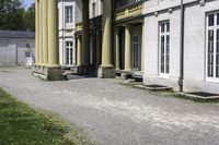 a building with columns is next to a walkway outside of it there are grass and stone in front of the entrance and steps