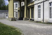 a building with columns is next to a walkway outside of it there are grass and stone in front of the entrance and steps