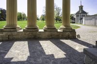 several pillars, in shadow, on a sidewalk in a grassy park area with a lawn and buildings