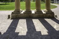 several pillars, in shadow, on a sidewalk in a grassy park area with a lawn and buildings