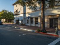 a white building on the side of the road with a sign on the sidewalk and bushes