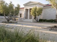 two trees and benches sit outside a building with columns and pillars on the outside, grass and bushes