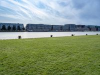 a grassy field in front of tall buildings and a lake with a few trees on top