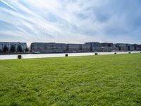 a grassy field in front of tall buildings and a lake with a few trees on top