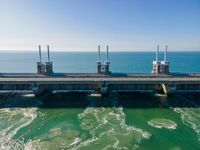 a bridge over the ocean with construction equipment in the background and two people walking on it