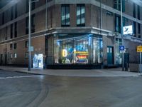 a lighted store sits outside a brown brick building in a city at night with no traffic