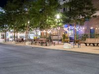 a empty sidewalk and street lined with benches and tables in a city at night time