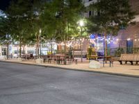 a empty sidewalk and street lined with benches and tables in a city at night time