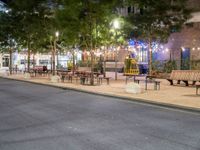 a empty sidewalk and street lined with benches and tables in a city at night time