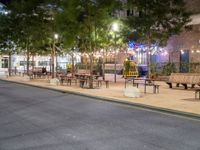 a empty sidewalk and street lined with benches and tables in a city at night time