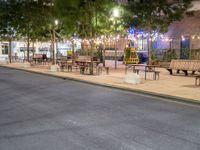 a empty sidewalk and street lined with benches and tables in a city at night time