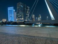 a woman is walking down the sidewalk by the water at night with city lights in the background