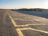 yellow lines on asphalt by sand and hills during sunrise as a vehicle travels away from a car