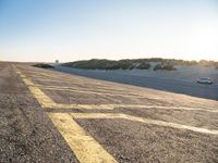 yellow lines on asphalt by sand and hills during sunrise as a vehicle travels away from a car