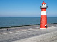 a red and white lighthouse on the beach with people walking up it to the lighthouse