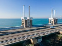 two cars driving on a large bridge over the water by sea level with wind turbines behind them