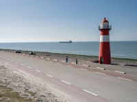 the lighthouse is by the water with boats out on the shore and people walking near the shore