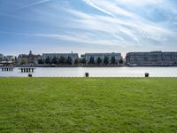 an empty park with the water in front of some buildings and a green field under a blue sky