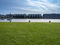 an empty park with the water in front of some buildings and a green field under a blue sky