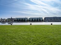 an empty park with the water in front of some buildings and a green field under a blue sky