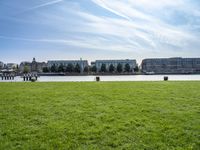 an empty park with the water in front of some buildings and a green field under a blue sky