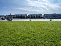 an empty park with the water in front of some buildings and a green field under a blue sky