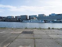 an empty concrete sidewalk near the water with a boat in the distance behind it and city buildings in the back