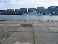 an empty concrete sidewalk near the water with a boat in the distance behind it and city buildings in the back