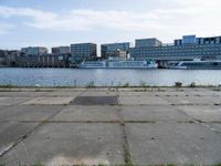 an empty concrete sidewalk near the water with a boat in the distance behind it and city buildings in the back