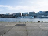 an empty concrete sidewalk near the water with a boat in the distance behind it and city buildings in the back