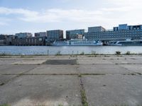 an empty concrete sidewalk near the water with a boat in the distance behind it and city buildings in the back