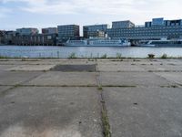 an empty concrete sidewalk near the water with a boat in the distance behind it and city buildings in the back