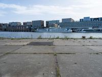 an empty concrete sidewalk near the water with a boat in the distance behind it and city buildings in the back