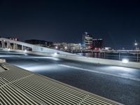 an empty city street with a bench on it at night near a river with the city lights reflecting off the water