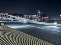 an empty city street with a bench on it at night near a river with the city lights reflecting off the water