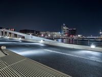 an empty city street with a bench on it at night near a river with the city lights reflecting off the water