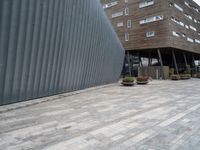 brick walkway next to an industrial building with wood trimming on the wall, and grey cement patio area, near planters and concrete sidewalk area with square seating area