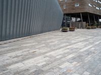 brick walkway next to an industrial building with wood trimming on the wall, and grey cement patio area, near planters and concrete sidewalk area with square seating area