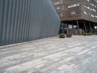 brick walkway next to an industrial building with wood trimming on the wall, and grey cement patio area, near planters and concrete sidewalk area with square seating area